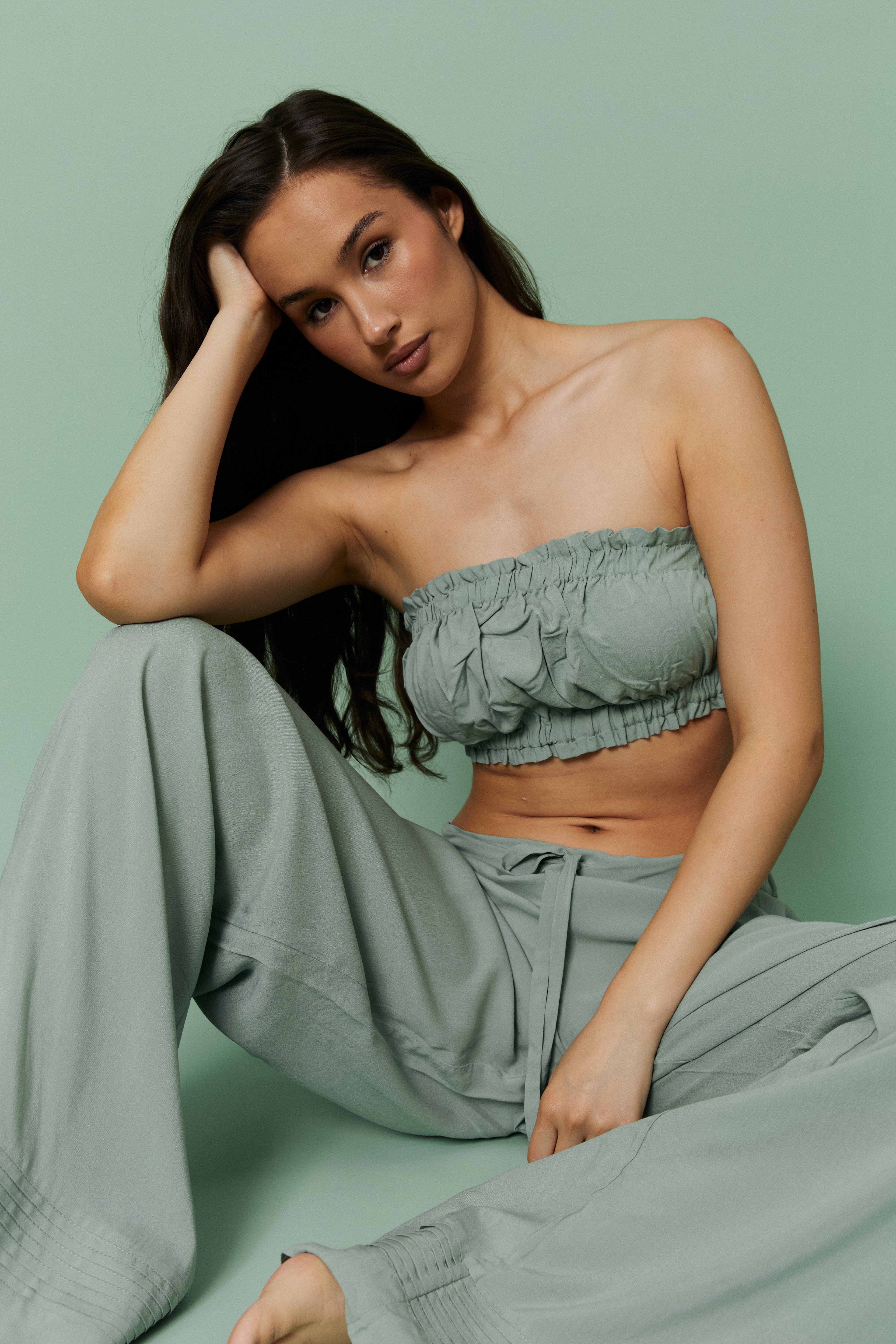 Seated model wearing Azalea Green Voile Crop top and matching pants, hand resting on head, highlighting ruched bandeau neckline and lightweight sheer voile fabric.