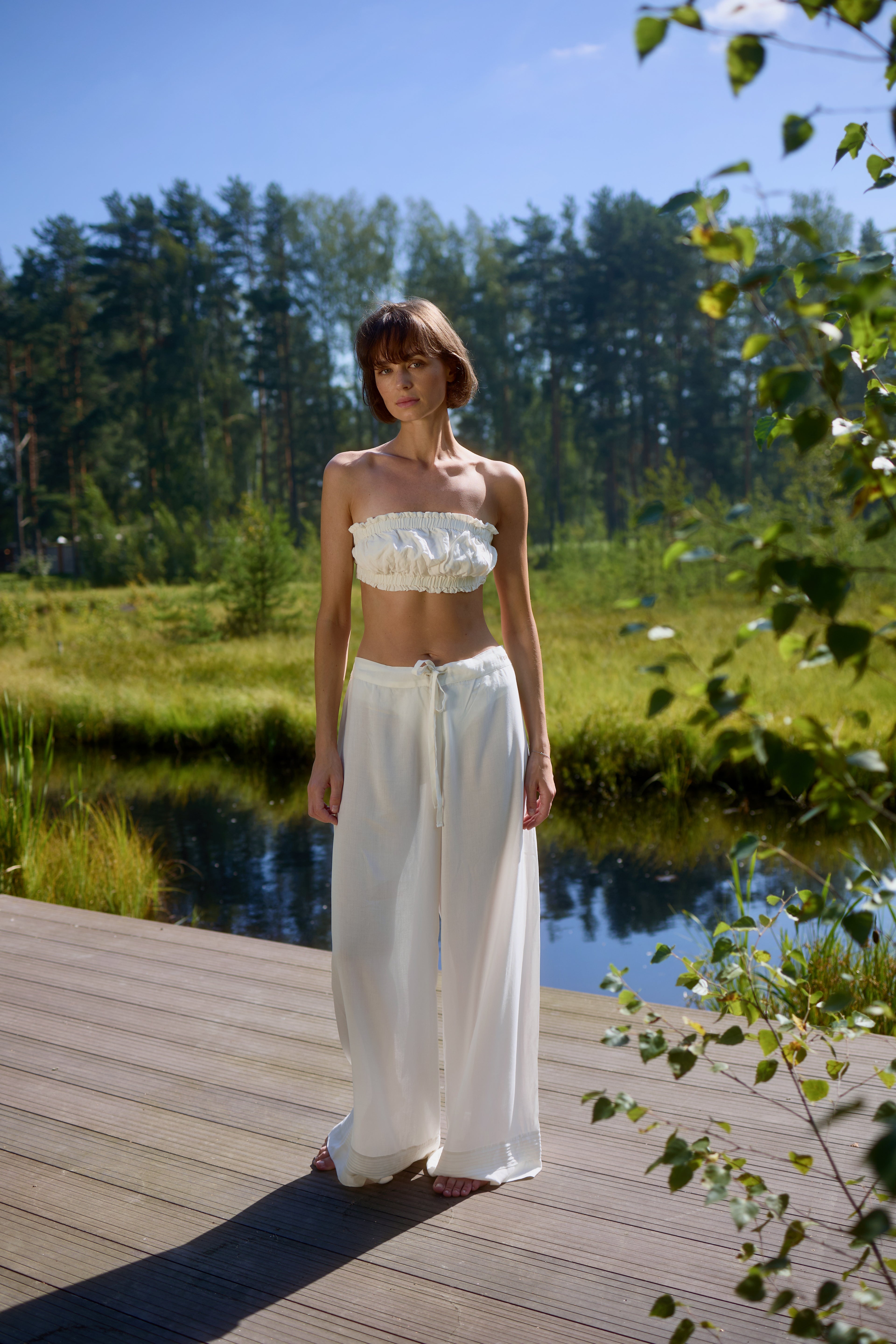 Full-length view of model standing on wooden deck by the lake wearing Azalea White Voile Lounge Bottom with matching crop top, showcasing soft flowing drape and relaxed luxury loungewear in natural sunlight.