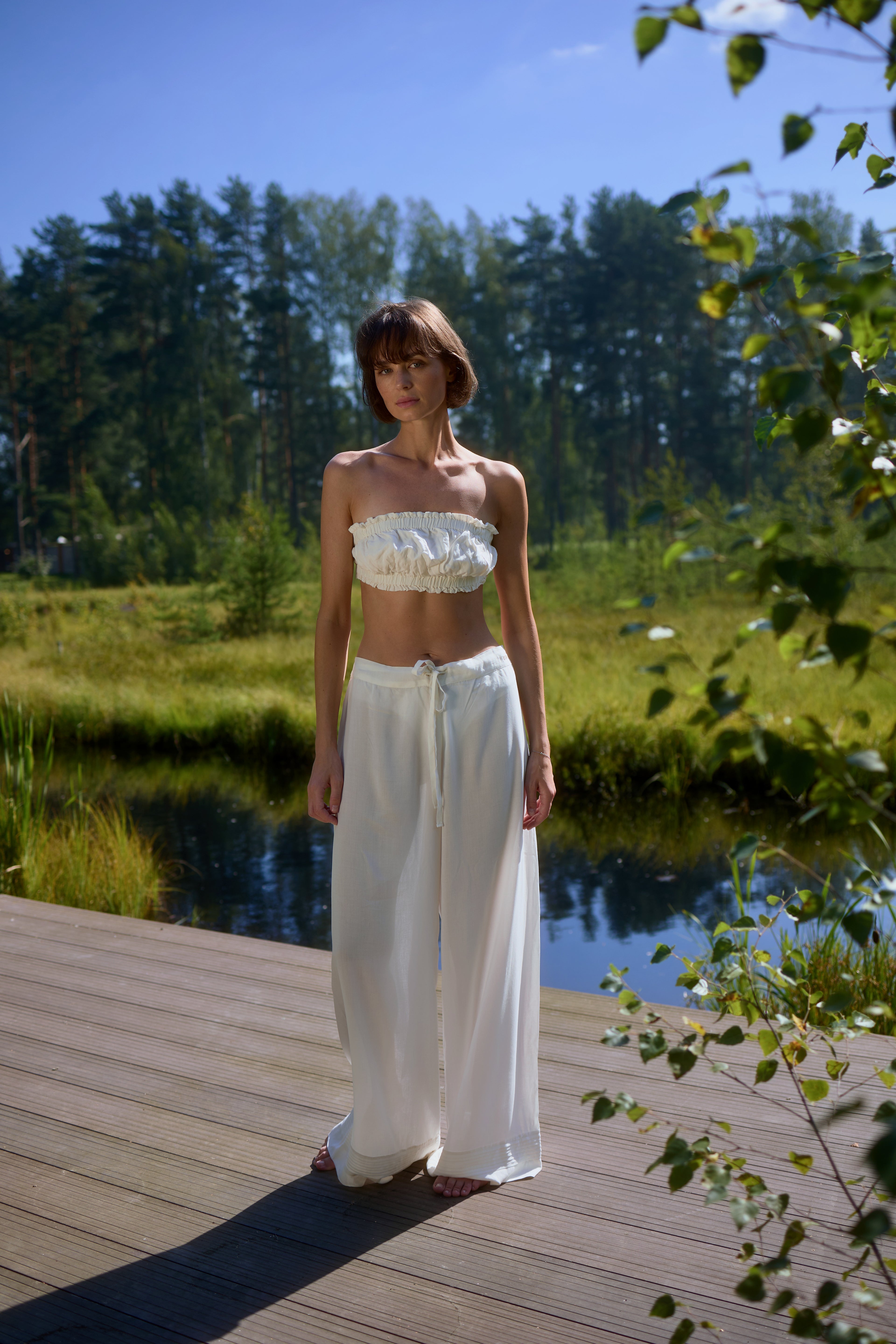 Full-length view of model standing on wooden deck by the lake wearing Azalea White Voile Lounge Bottom with matching crop top, showcasing soft flowing drape and relaxed luxury loungewear in natural sunlight.