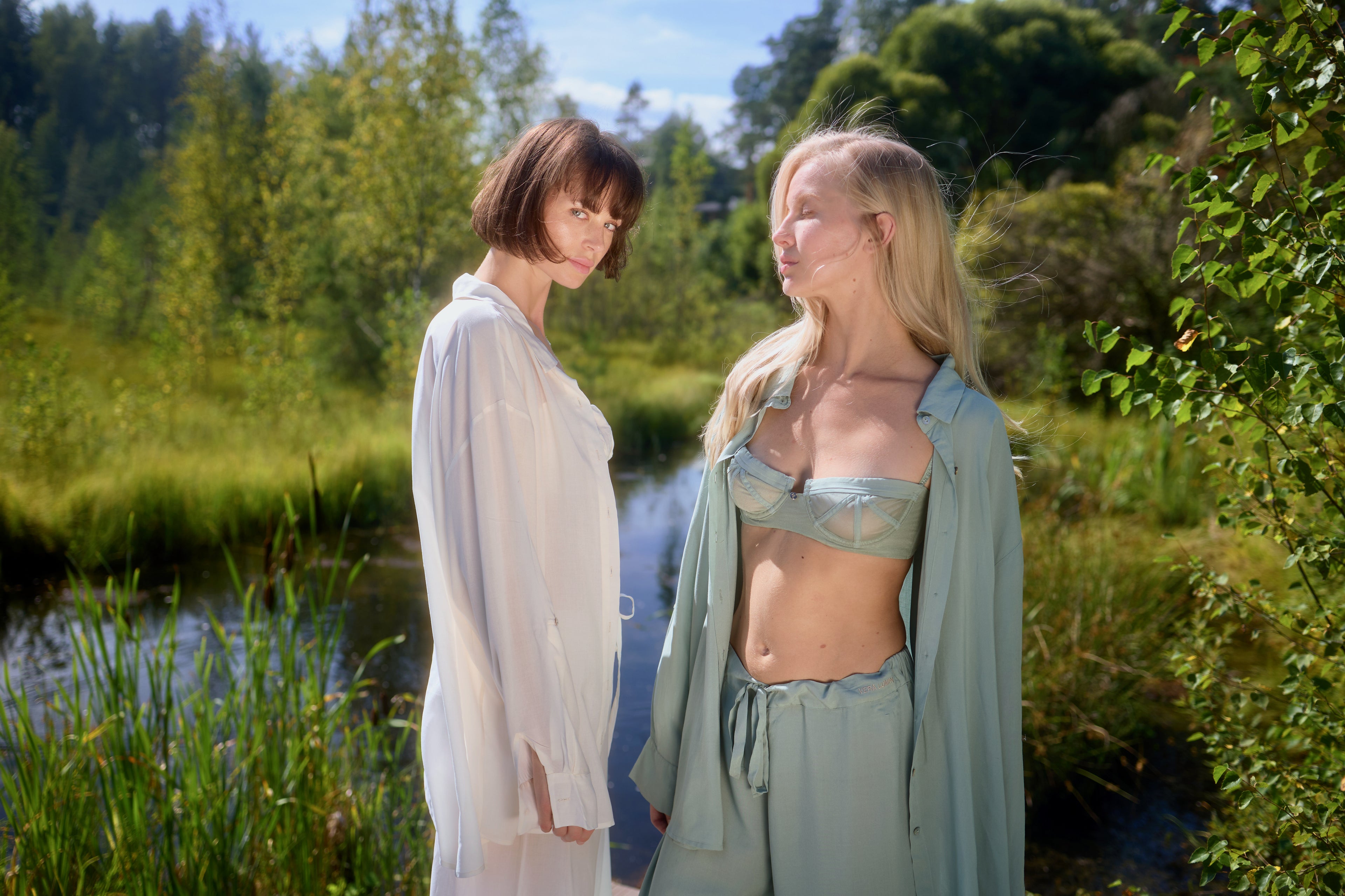 Two models standing by a sunny lake in Azalea loungewear: brunette in white voile shirt, blonde in green voile balconette bra and lounge bottom with open shirt.