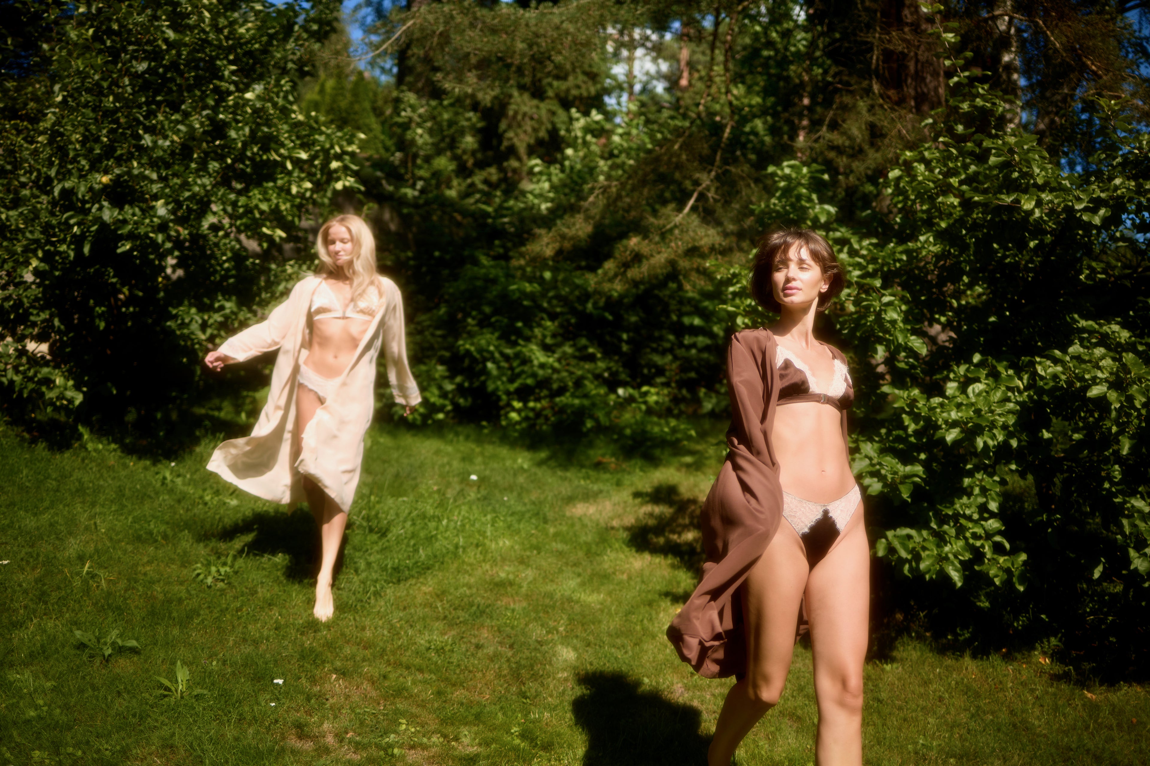 Two models in garden wearing Monsella Silk Robes and lingerie: Powder Silk Robe with Powder Triangle Bra and Slip on the left, Brown Silk Robe with Brown Triangle Bra and Slip on the right.