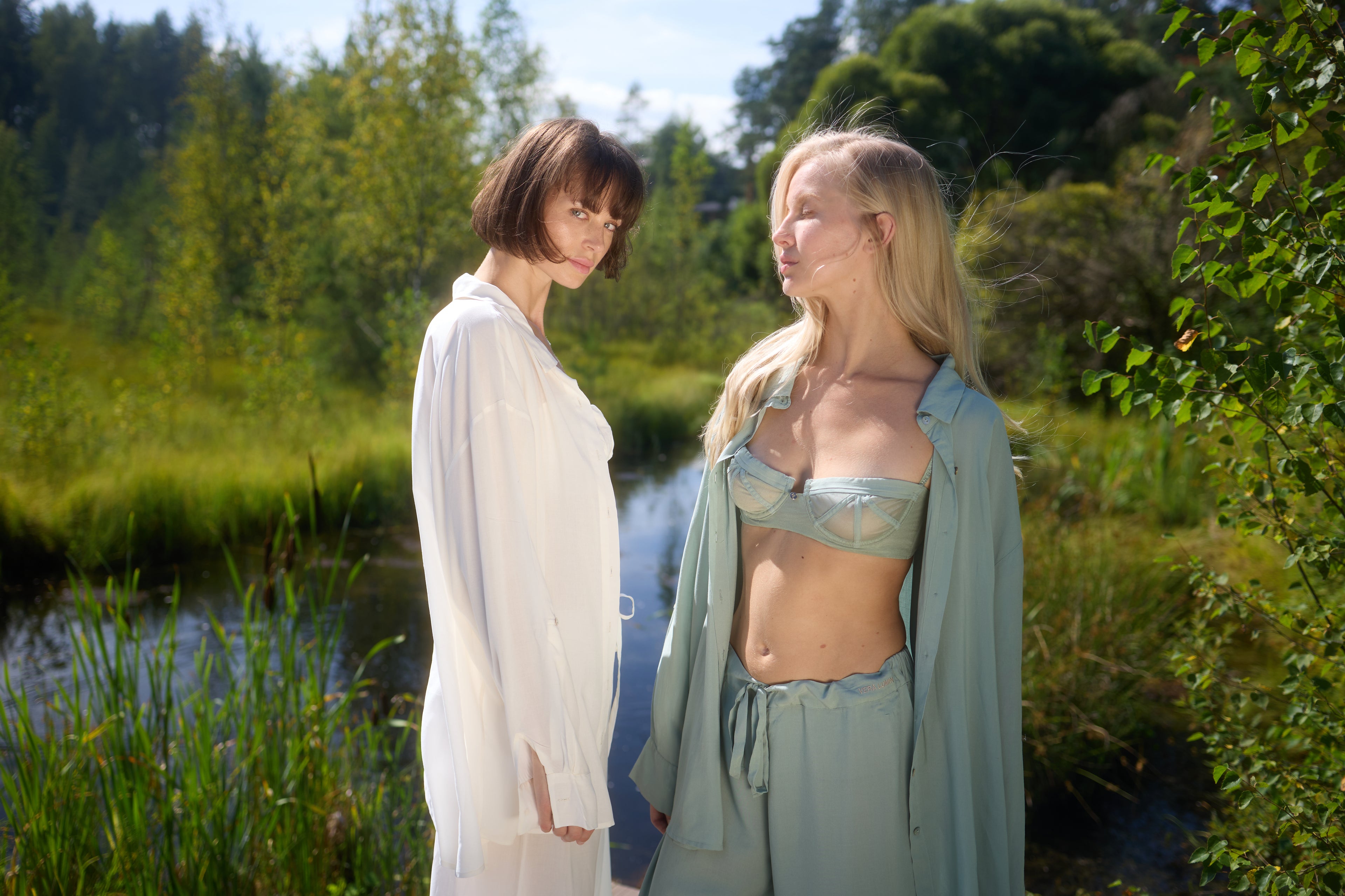 Two models standing by a sunny lake in Azalea loungewear: brunette in white voile shirt, blonde in green voile balconette bra and lounge bottom with open shirt.