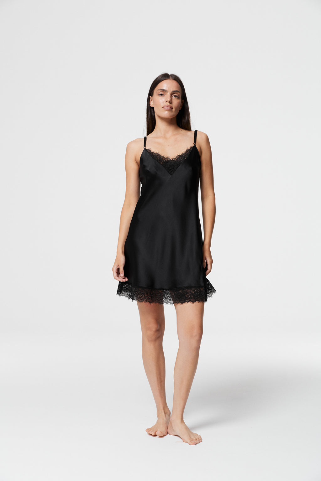 Model standing in full frontal view wearing Dicentra Black Silk Nightdress, featuring luxurious black silk fabric, delicate lace V-neckline, thin spaghetti straps and lace-trimmed hem.